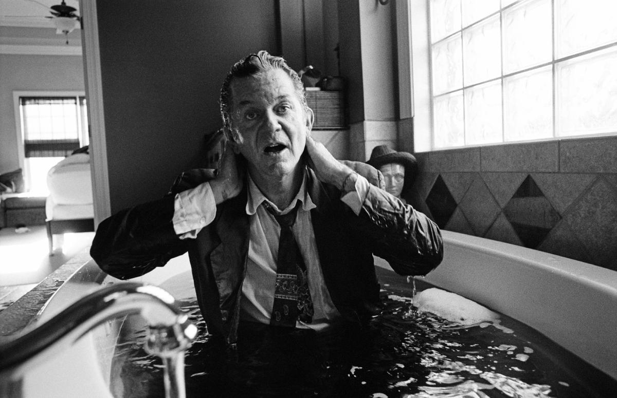 A man in a suit sits in a bathtub, hands to his head, mouth open in a shout.